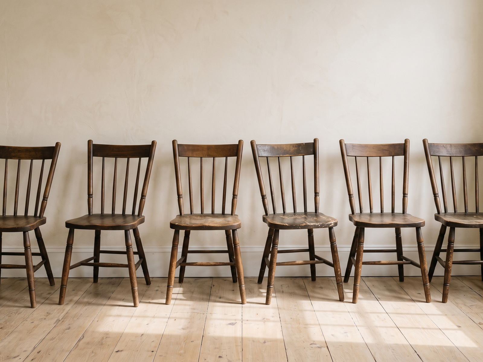 A row of six wooden chairs standing against a plaster wall, each one at a slightly different angle.