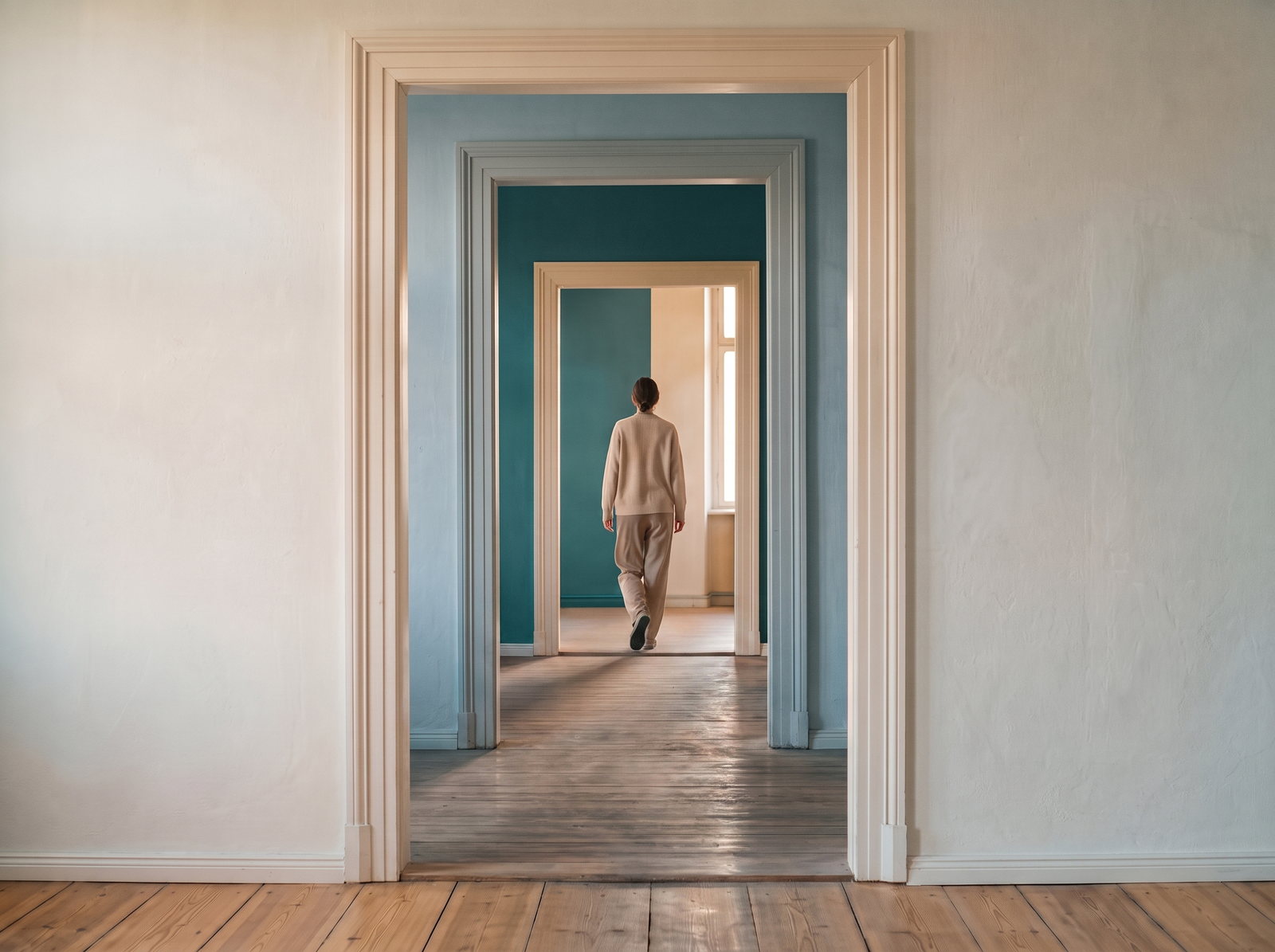 A person in soft neutral clothing walks away through a series of open doorways toward a sunlit room, where morning light curves across the wooden floor.
